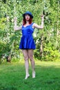 Woman on the lawn with red curly hair in a blue hat and short blue mini dress. Hands raised up and bent at the elbows.