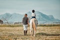 .Woman leading child on horse, ranch and mountain in background lady and animal walking on field from back. Countryside Royalty Free Stock Photo