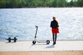 Woman in leather jacket stands near electric scooter on sunny promenade backside view