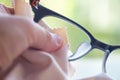 Woman left hand cleaning grey shortsighted or nearsighted eyeglasses by beige microfibre cleaning cloths, Bokeh green bg.