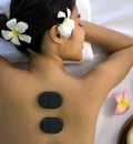 A woman lies on a massage table, her back exposed, as a therapist uses heated stones to deliver a soothing back massage. The hot