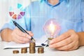 Woman with light bulb, notebook and coins at table, closeup. Energy saving Royalty Free Stock Photo