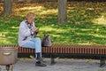 A woman in a light jacket sits on a bench and dials a number on a cell phone.