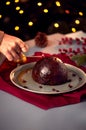 Woman Lighting Brandy Soaked Christmas Pudding On Table Set For Festive Christmas Meal