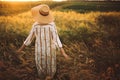 Woman in linen dress and hat walking in wildflowers and herbs in sunset golden light in summer meadow. Stylish rustic girl Royalty Free Stock Photo