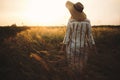 Woman in linen dress and hat walking in wildflowers and herbs in sunset golden light in summer meadow. Stylish rustic girl Royalty Free Stock Photo