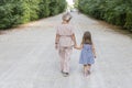 A woman and a little girl walking on a dirt road along a forest under sunny sky, back view from behind.