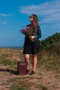 Woman with long brown hair in denim jacket, black skirt, vintage suitcase, flowers bouquet off-road. Lifestyle photo Royalty Free Stock Photo