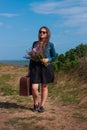 Woman with long brown hair in denim jacket, black skirt, vintage suitcase, flowers bouquet off-road. Lifestyle photo Royalty Free Stock Photo