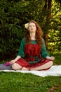 A woman with long curly red hair and closed eyes is sitting serenely on the grass in the lotus position. Meditations in nature