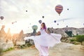 Woman in a long dress on background of balloons in Cappadocia. Girl with flowers hands stands on a hill and looks