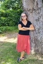 Woman with long grey hair and glasses and organge skirt and black blouse leans against large tree with arms crossed and smile on h Royalty Free Stock Photo