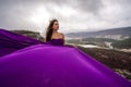 A woman with long hair is standing in a purple flowing dress with a flowing fabric. On the mountain against the Royalty Free Stock Photo