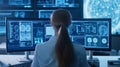A woman with long hair, wearing a lab coat, sits in front of multiple computer monitors displaying brain scans and data