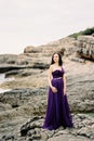 Woman in a long lilac dress stands on the rocks near the water