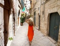 Woman with long orange summer dress walking in narrow alley of Bisceglie medieval historical town of Apulia, Italy