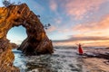 Woman in long red dress standing in ocean waves by a sea arch