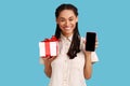 Woman looking at camera with toothy smile, showing present box and smart phone with blank screen. Royalty Free Stock Photo