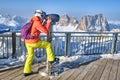 Woman looking through coin operated binoculars, with Sassolungo peaks in the background, at Terrazza Delle Dolomiti viewpoint