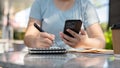 A woman looking at her smartphone and taking notes in her notebook at a table outdoors Royalty Free Stock Photo