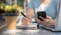 A woman looking at her smartphone and taking notes in her notebook at a table outdoors Royalty Free Stock Photo