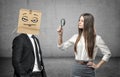 Woman is looking through a magnifier at a businessman with a box on his head with a pokerface