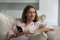 Woman looks aside sits on sofa with smartphone in hands