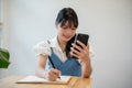 A woman making lists in her notebook while using her smartphone, sitting at a table indoors Royalty Free Stock Photo