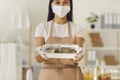 Woman in a mask gives the client a ready fresh healthy lunch in a plastic container.