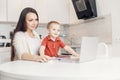 Woman on maternity leave working home online with laptop in kitchen with small child. Concept mom work while in Royalty Free Stock Photo