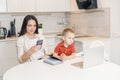 Woman on maternity leave working home online with laptop in kitchen with small child. Concept mom work while in Royalty Free Stock Photo