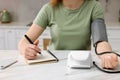 Woman measuring blood pressure and writing it down into notebook in kitchen, closeup Royalty Free Stock Photo