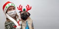 Woman in medical mask and Christmas hat with pug dog in traditional red Christmas deer. Coronavirus and Christmas concept. pet Royalty Free Stock Photo