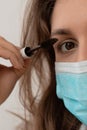 Woman in a medical mask does makeup, mascaras her eyes. The face of a young beautiful woman in a stylish mask