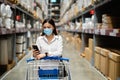 Woman with medical mask looking at her mobile phone and shopping in warehouse store during coronavirus covid-19 pandemic