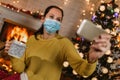 Woman in medical mask make selfie, sitting in front of decorated christmas tree and fireplace at home