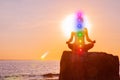 Woman is meditating with glowing seven chakras on stone at sunset. Silhouette of woman is practicing yoga on the beach