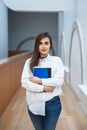 Woman model with long dark hair in white shirt and blue jeans standing in hall at college university holding notebook