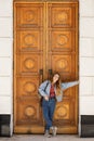 A woman in a modern denim outfit stands at closed massive high wooden doors with a classic pattern.
