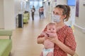 Woman mother in medical face mask holds baby boy in hospital lobby. Mom with child son in her arms visit in the clinic hall Royalty Free Stock Photo