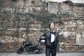 Woman a motorcyclist standing with helmet in hand near her bike, brick wall of garage background. Girl in a leather