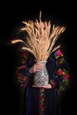 Woman in national dress holding a bouquet of dry wheat in a vase. On black bakground. Face covered with wheat