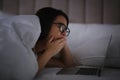 Young woman with nomophobia yawning while using laptop in bed at night. Insomnia concept