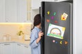 Woman opening refrigerator door with child`s drawing, notes and magnets