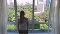 Woman opens curtain on the big windows city view with green gadern and buildings. Green city concept.