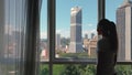 Woman opens curtain on the big windows city view with green gadern and buildings. Green city concept.