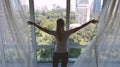 Woman opens curtain on the big windows city view with green gadern and buildings. Green city concept.