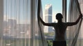 Woman opens curtain on the big windows city view with green gadern and buildings. Green city concept.