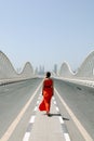 A woman in an orange red dress walking by an empty road on a Meydan bridge with city view on background in Dubai
