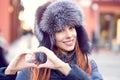 Woman outside in winter fur hat showing heart shape with hand.